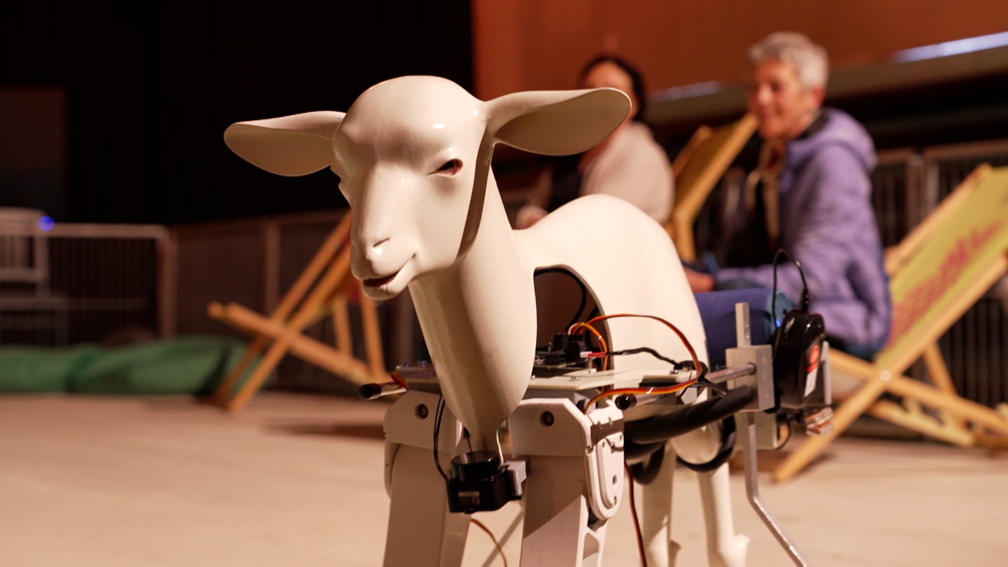 Eine weiße Roboterziege starrt leblos in die Kamera, Kabel hängen aus dem Körper heraus, im Hintergrund Zuschauer auf Liegestühlen. | Bild: BR/Simon Frick Eine weiße Roboterziege starrt leblos in die Kamera, Kabel hängen aus dem Körper heraus, im Hintergrund Zuschauer auf Liegestühlen.