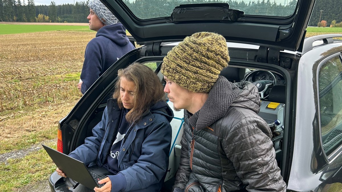 Ein Team von Labfly überwacht und steuert die Drohne, zwei Menschen sitzen mit Laptop im Kofferraum eines Autos und der Drohnenpilot lehnt am Auto und lenkt die Drohne per Joystick