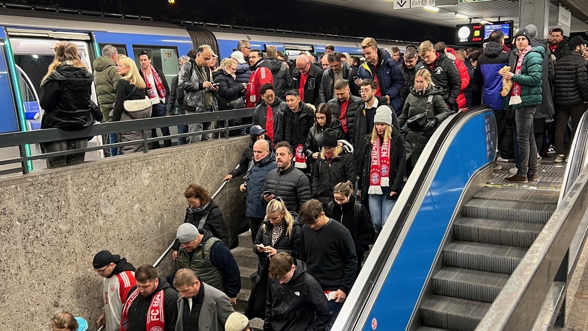 Ein U-Bahnsteig in München ist überfüllt mit Passagieren, von denen einige Fanschals und Trikots des FC Bayern tragen.