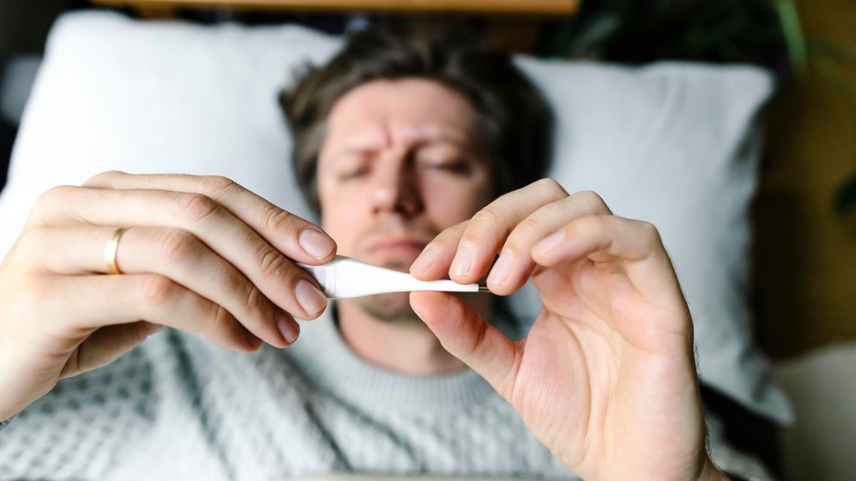 Erkältung, Grippe, Corona oder RS-Virus? Mann liegt im Bett und hält ein Fieberthermometer in der Hand.