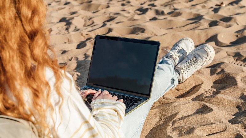 Eine Frau mit roten Haaren sitzt am Strand im Sand und hat einen Laptop auf den Beinen.