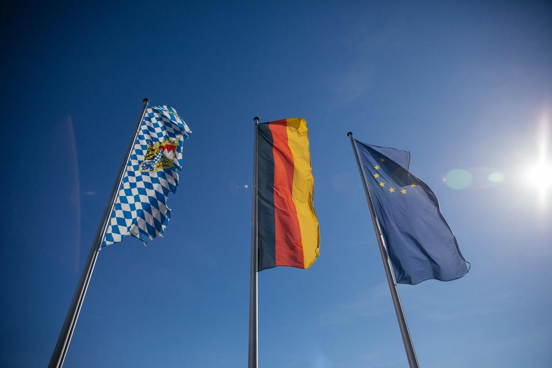 Drei Flaggen an Flaggenmasten aus der Froschperspektive: Von links nach rechts die weiß-blaue Rautenflagge Bayerns, die schwarz-rot-goldene Deutschlandflagge und die blaue Europaflagge mit gelbem Sternenr