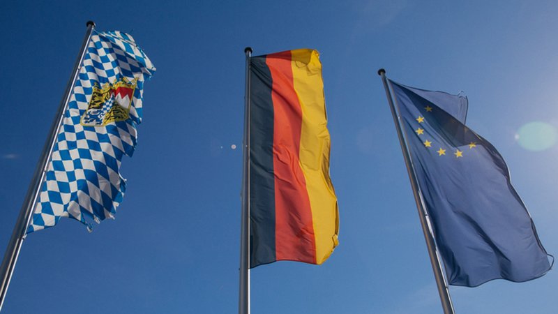 Drei Flaggen an Flaggenmasten aus der Froschperspektive: Von links nach rechts die weiß-blaue Rautenflagge Bayerns, die schwarz-rot-goldene Deutschlandflagge und die blaue Europaflagge mit gelbem Sternenr | Bild: BR Julia Müller Drei Flaggen an Flaggenmasten aus der Froschperspektive: Von links nach rechts die weiß-blaue Rautenflagge Bayerns, die schwarz-rot-goldene Deutschlandflagge und die blaue Europaflagge mit gelbem Sternenr