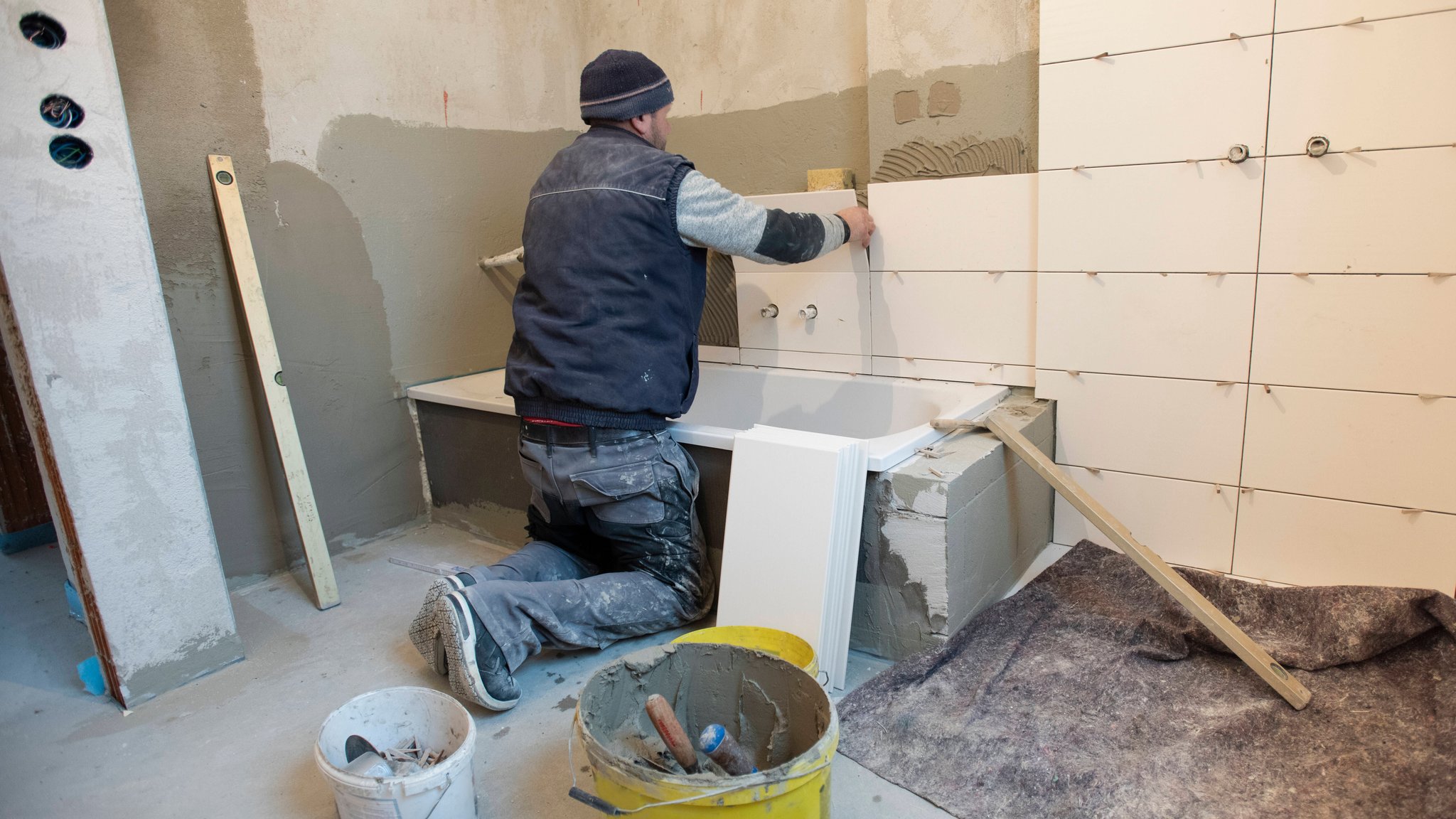 In einem Neubau klebt ein Bauarbeiter und Fliesenleger neue Fliesen an die Wand von einem Badezimmer.