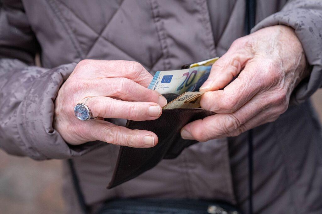 Die Hände einer älteren Person halten eine geöffnete Geldbörse, aus der ein 20-Euro-Schein hervorragt. 