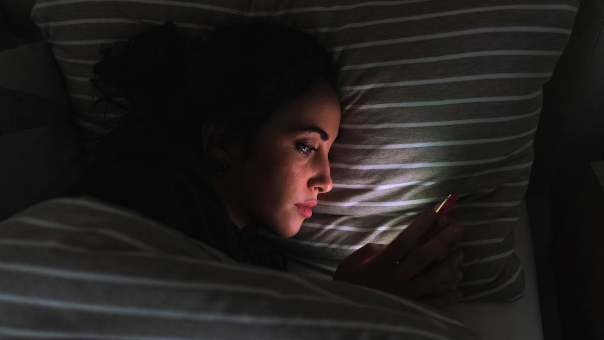 Eine Frau scrollt durch ihr Handy, liegt im Dunklen in ihrem Bett.