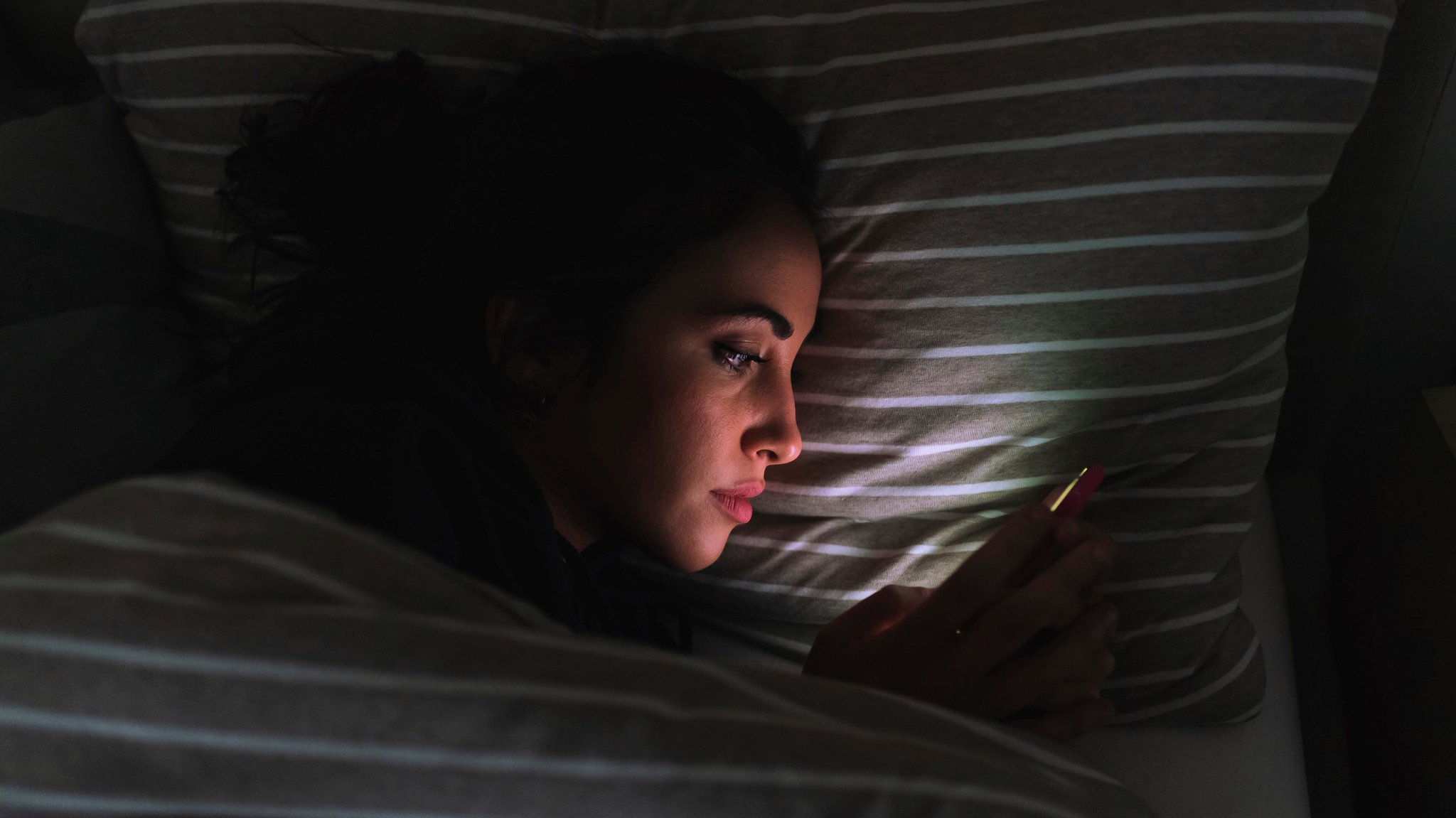 Eine Frau scrollt durch ihr Handy, liegt im Dunklen in ihrem Bett.