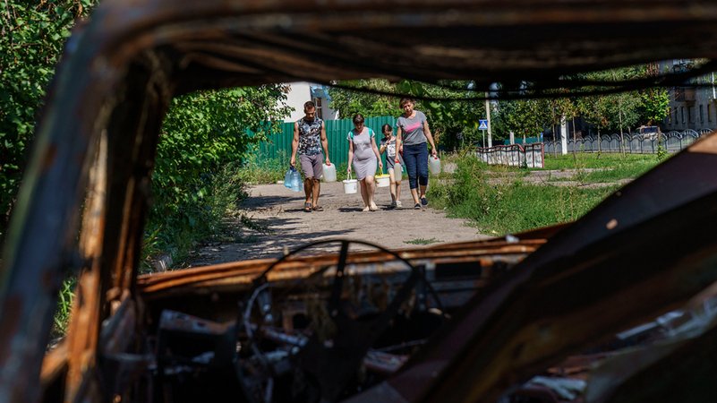 Anwohner tragen Behälter mit Wasser einen Weg in Richtung eines Autos, dass durch Beschuss des russischen Militärs zerstört wurde (Archivbild).