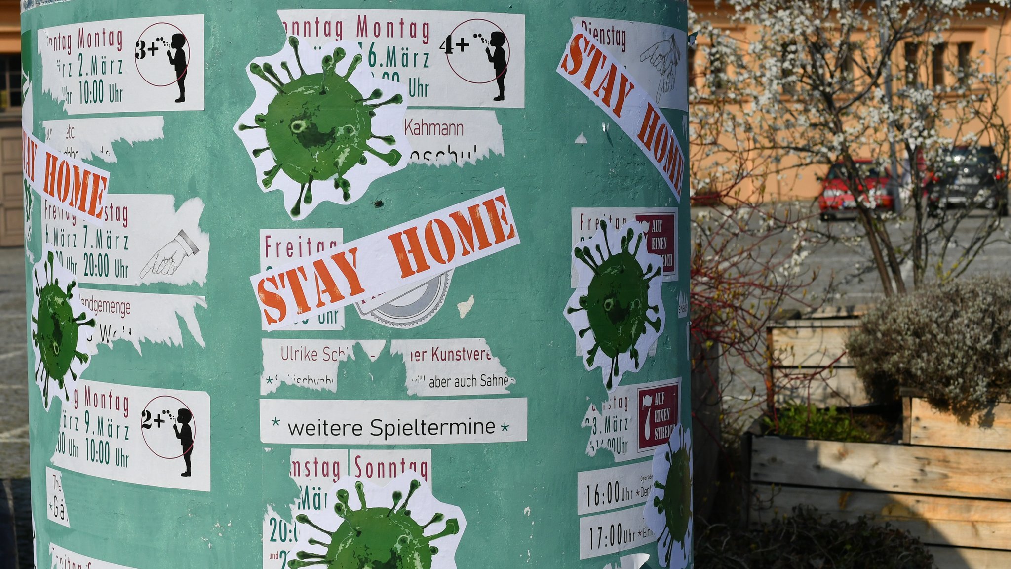 Litfaßsäule mit Veranstaltungen, die aufgrund von Corona abgesagt wurden. Darüber Aufkleber von stilisiertem Virus und der Aufforderung "STAY HOME".