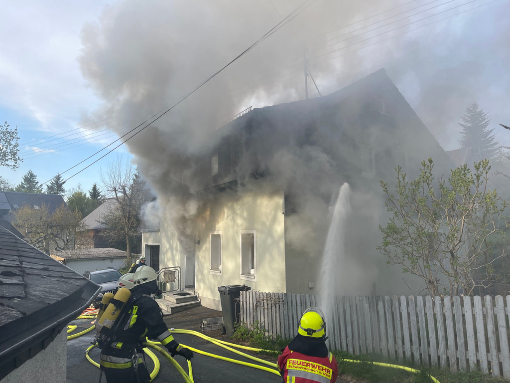 Feuerwehrleute mit Atemschutz löschen den Brand eines Hauses, aus dem dichte schwarze Rauchwolken dringen. 