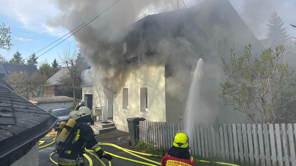 Feuerwehrleute mit Atemschutz löschen den Brand eines Hauses, aus dem dichte schwarze Rauchwolken dringen. 