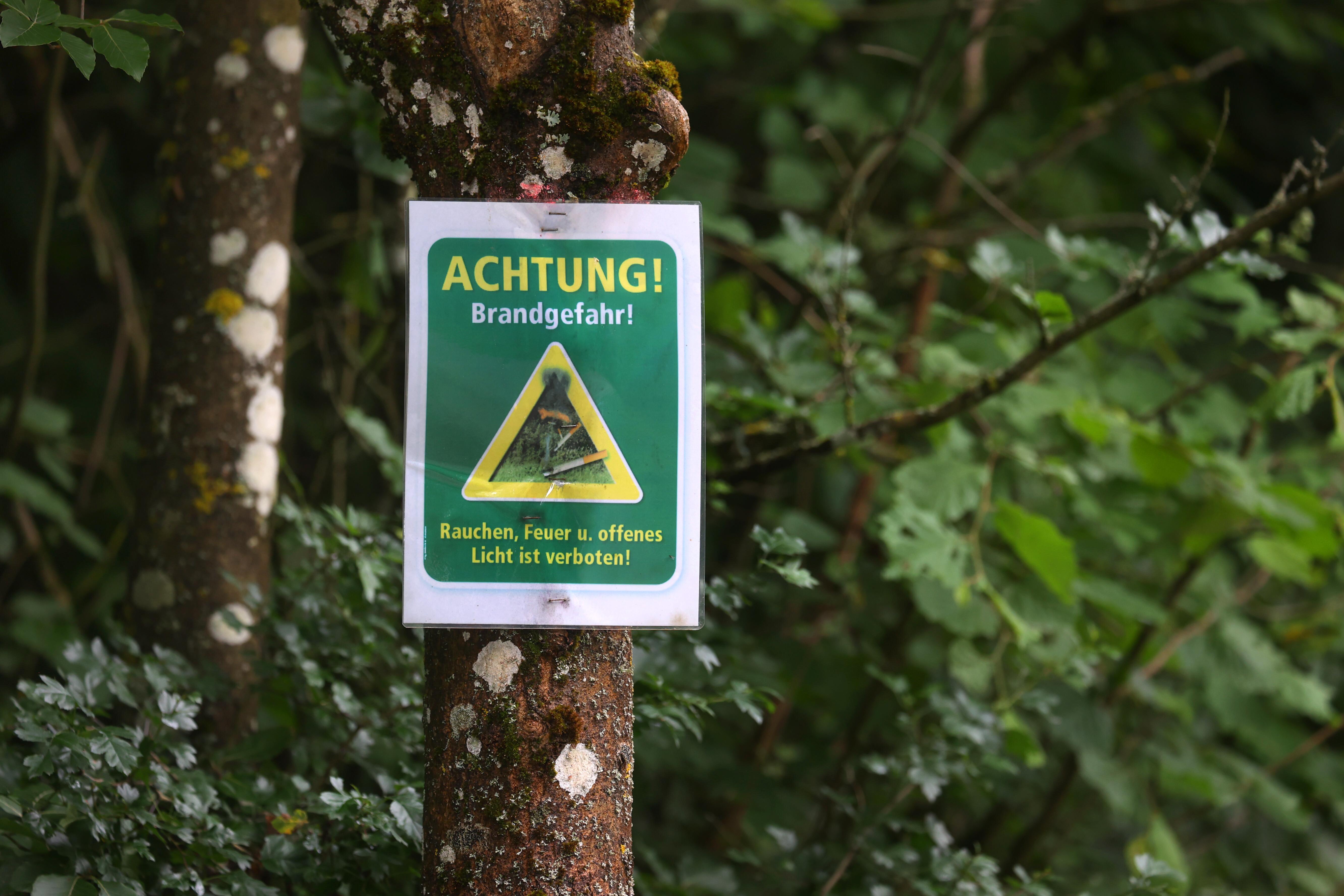 (Symbolbild) Ein Schild mit der Aufschrift "Achtung! Brandgefahr! - Rauchen, Feuer und offenes Licht ist verboten" hängt in einem Wald an einem Baum.