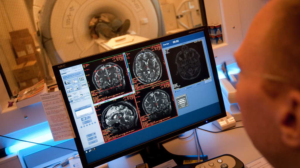Ein Laborant sitzt in der Uniklinik Großhadern in München (Oberbayern) vor einem Bildschirm mit Aufnahmen eines MRT vom Gehirn des Probanden, der im Hintergrund im MRT-Gerät liegt. 