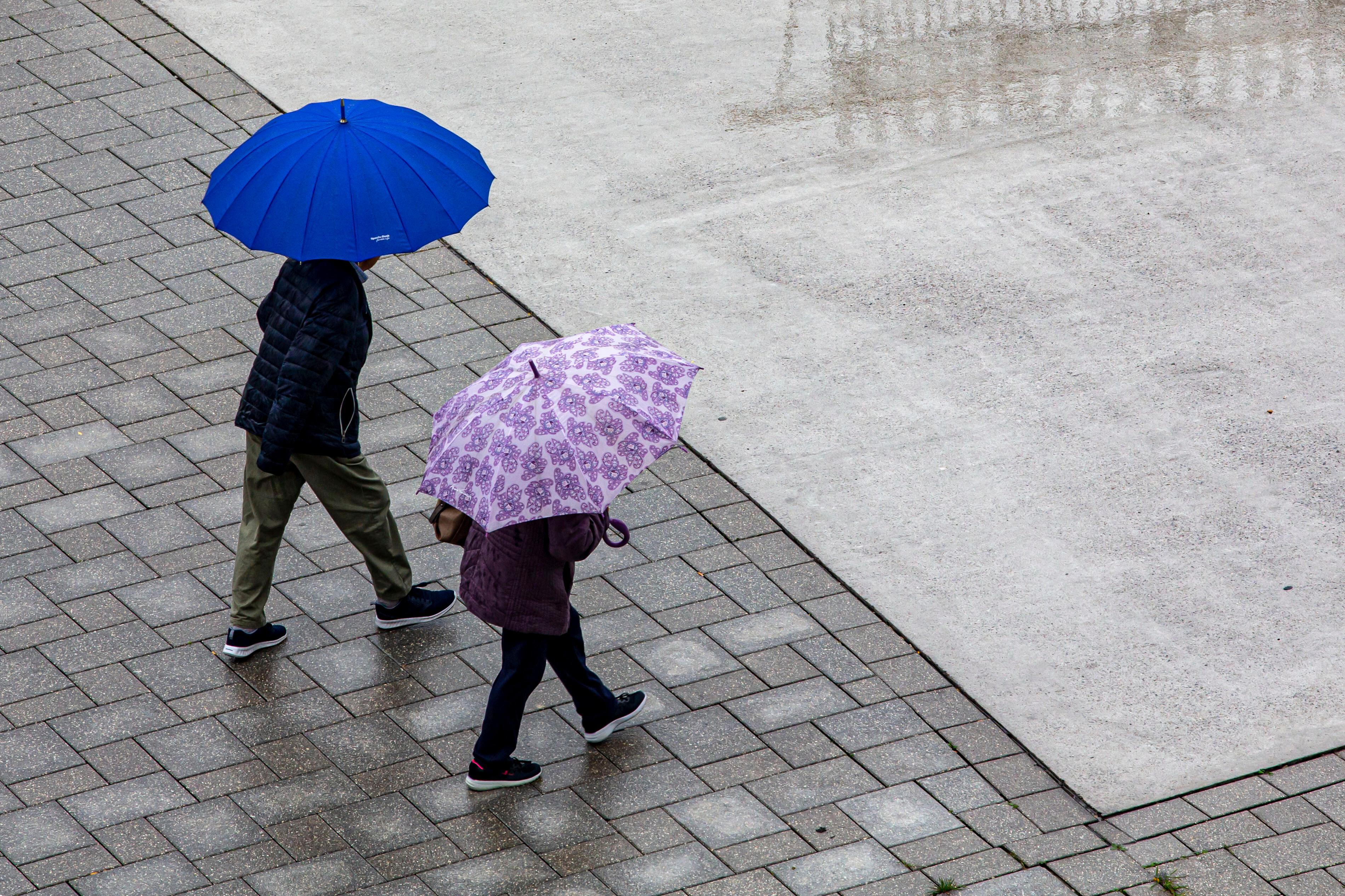 Zwei Menschen mit Regenschirmen gehen auf einem Platz nebeneinander. 