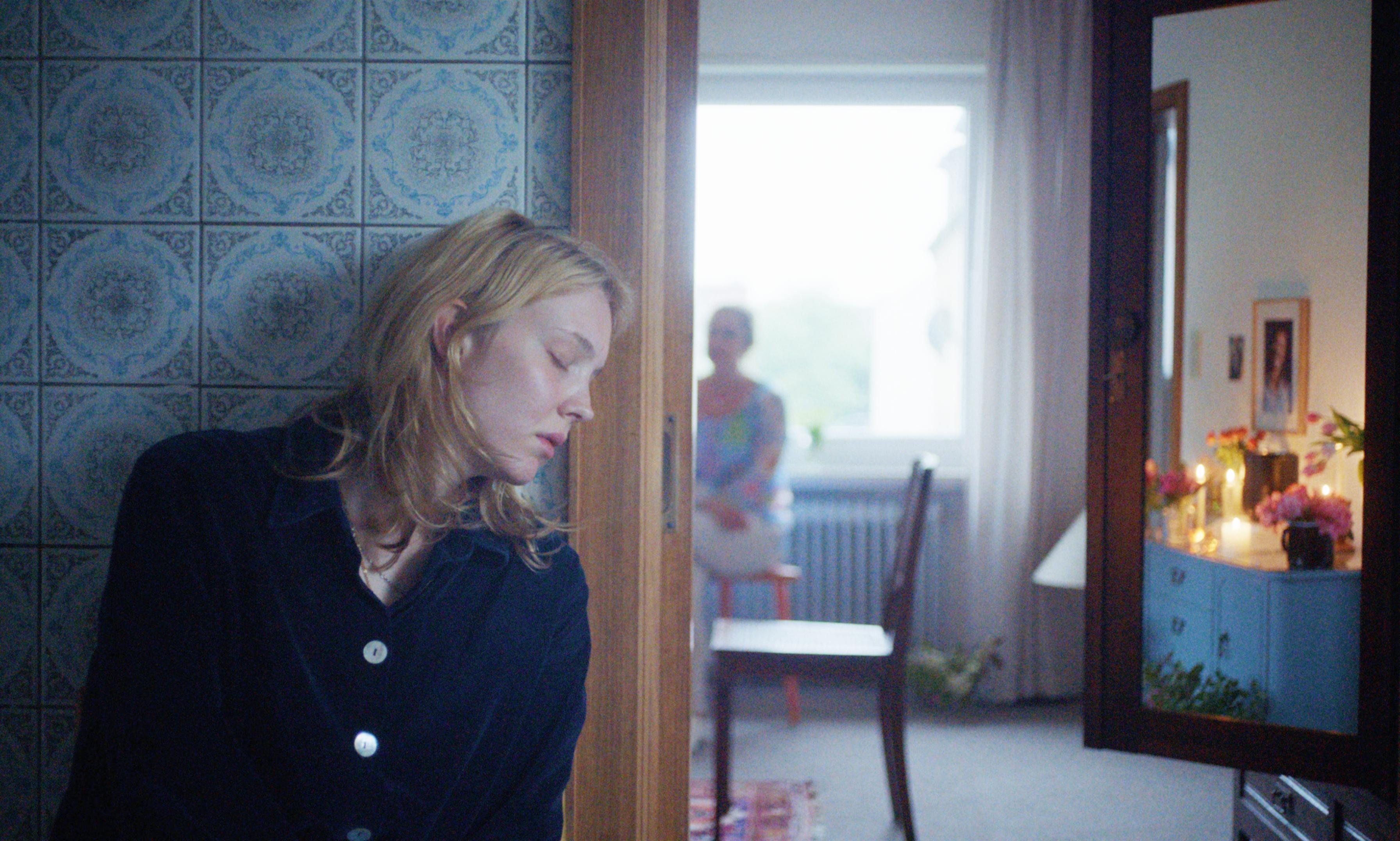 Eine junge Frau lehnt mit geschlossenen Augen an einer gekachelten Wand, im Hintergrund ist ein Traueraltar in einem Esszimmer zu erkennen. | Bild:Filmweh / Markus Ott