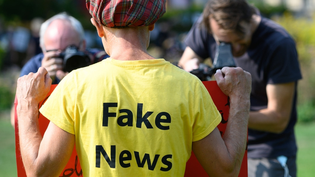 Bei einem Protest gegen Corona-Maßnahmen in Dresden hält ein Mann mit karierter Mütze und gelbem T-Shirt mit der Rückseiten-Aufschrift "Fake News" ein rotes Plakat in Richtung zweier Fotografen.