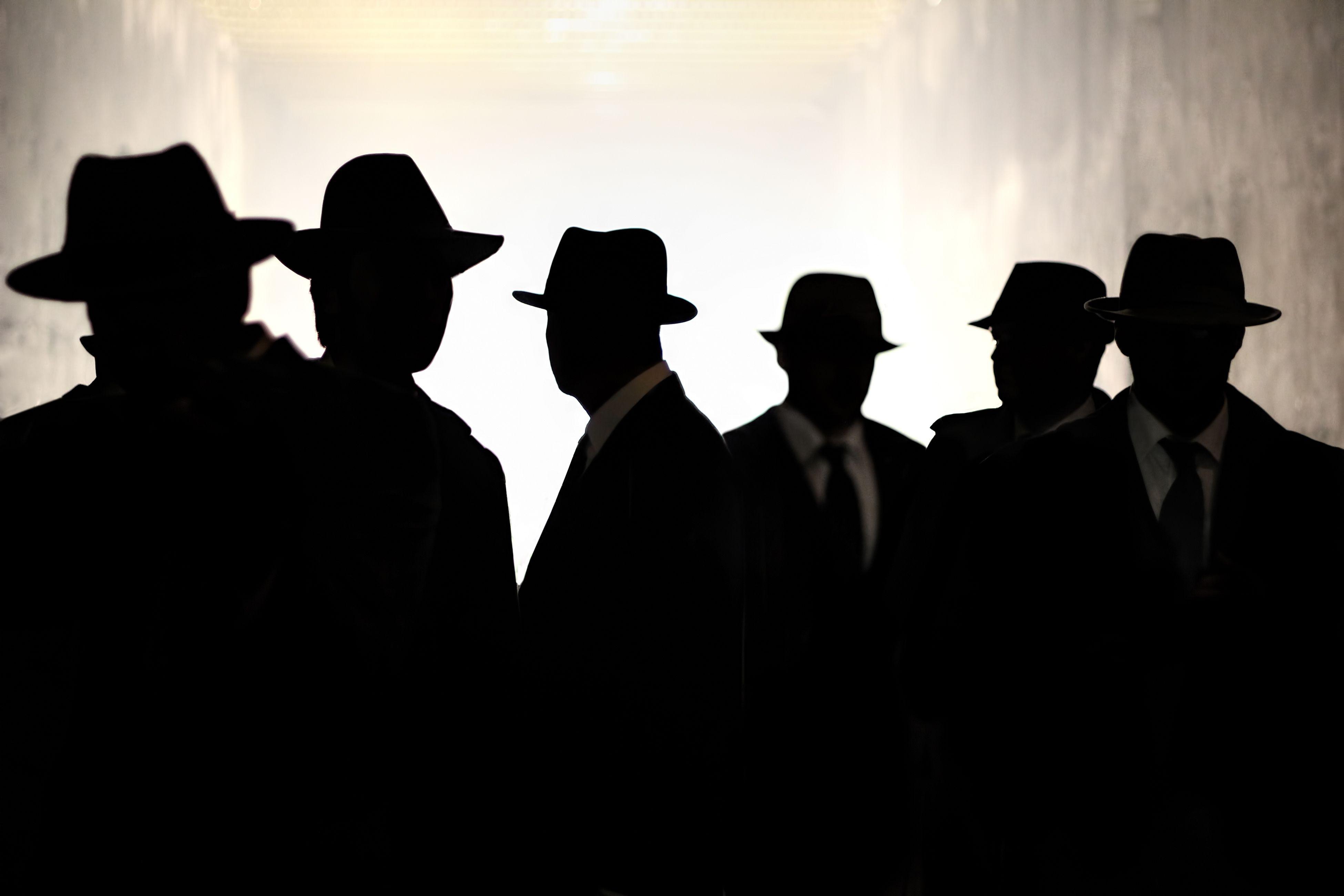 Silhouetten von Männergestalten, die nicht zu erkennen sind. Sie tragen schwarze Anzüge und Hüte