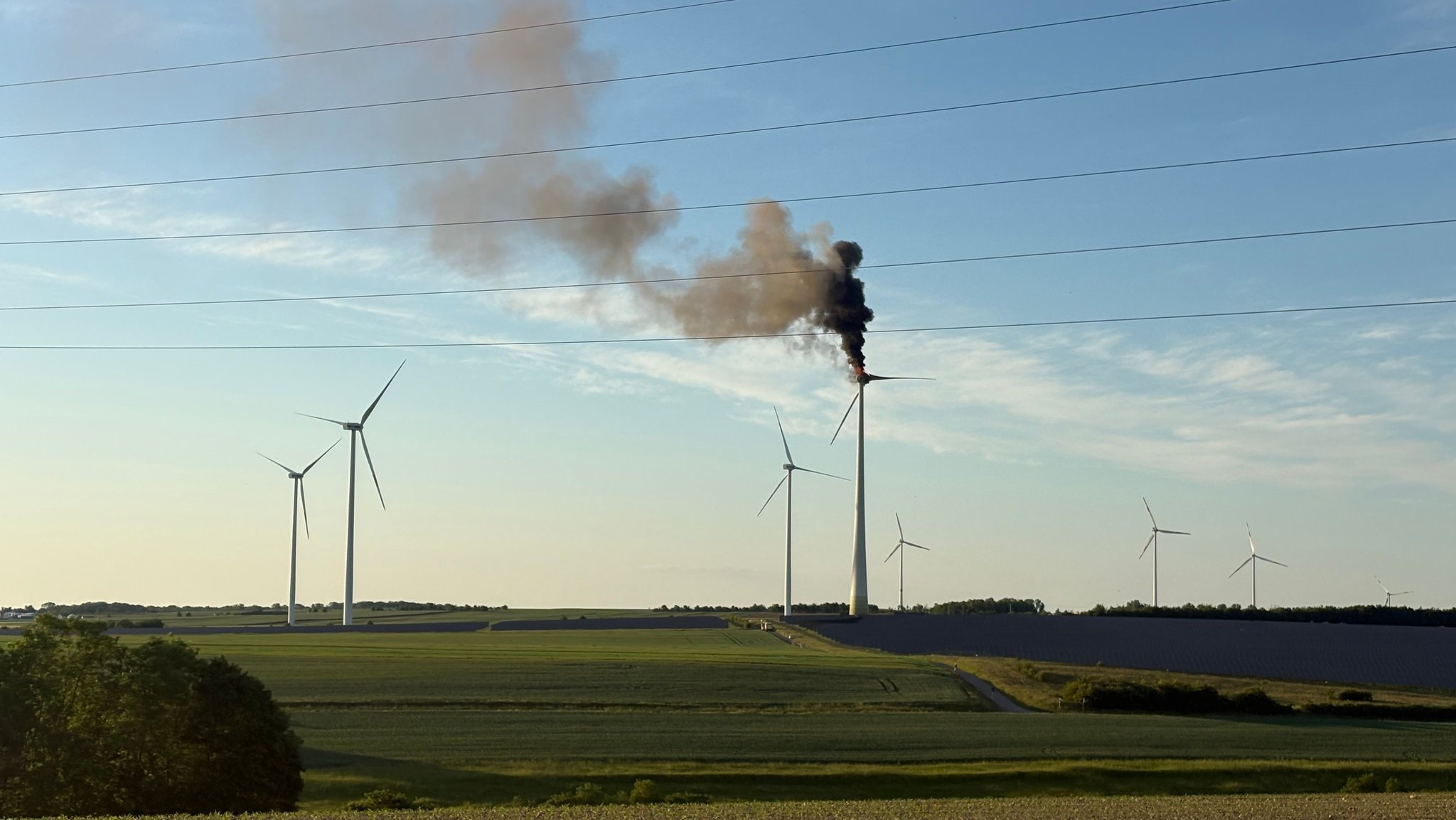 Mehrere Windräder auf einem freien Feld, von dem eines brennt. Schwarzer Rauch steigt von ihm den Himmel hoch.