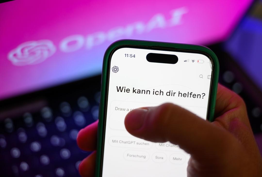 Ein Smartphone mit dem KI-Chatbot ChatGPT ist vor dem Logo des US-Softwareunternehmens OpenAI zu sehen.