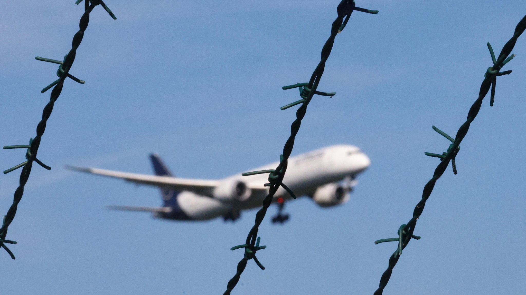 Symbolbild: Ein Flugzeug im Landeanflug, im Vordergrund des Bildes ist Stacheldraht zu sehen.