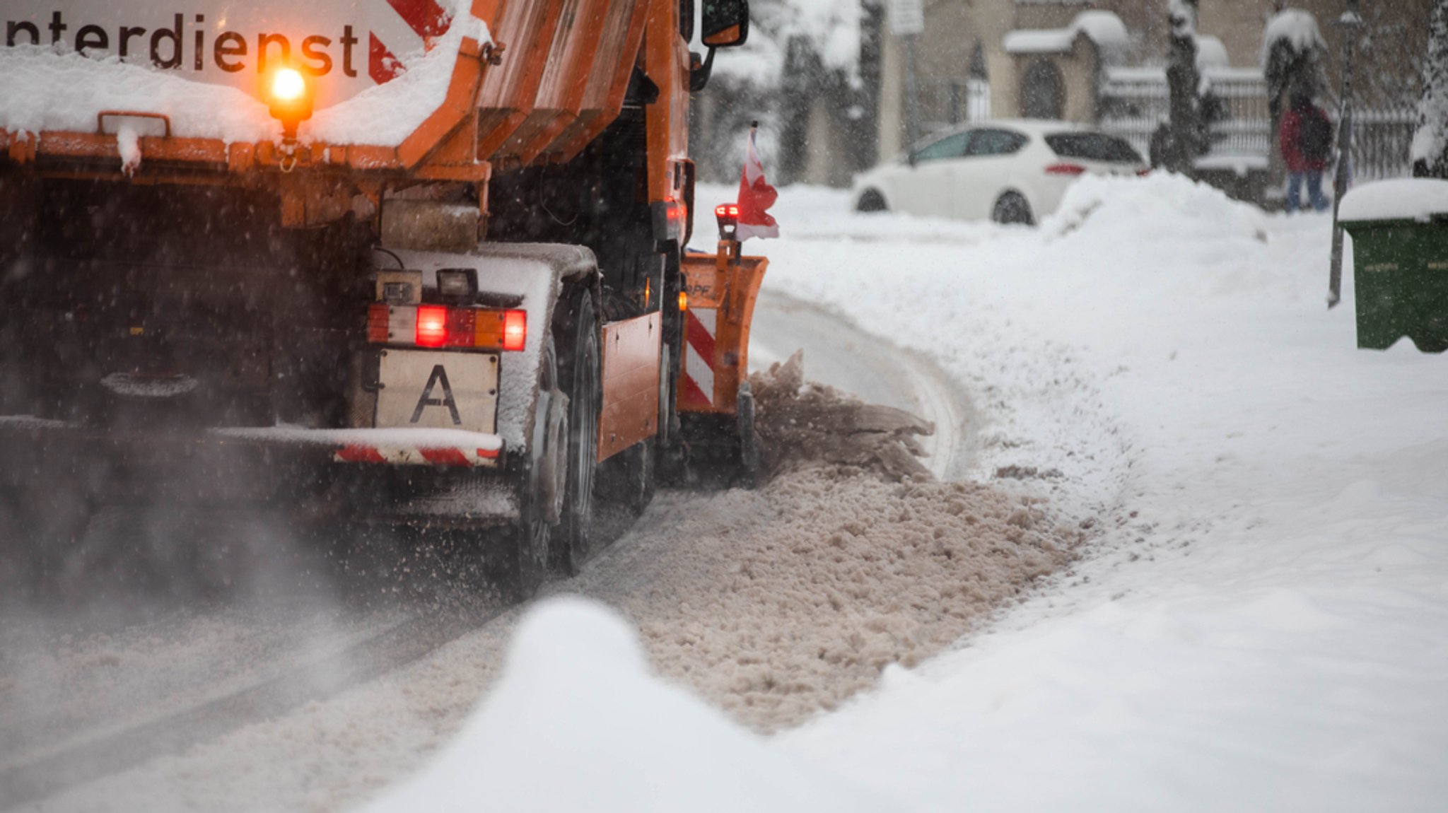 Ein Schneepflug des Winterdienstes von hinten; man sieht die orangene Warnleuchte und den Streuer im Betrieb. (Symbolbild)