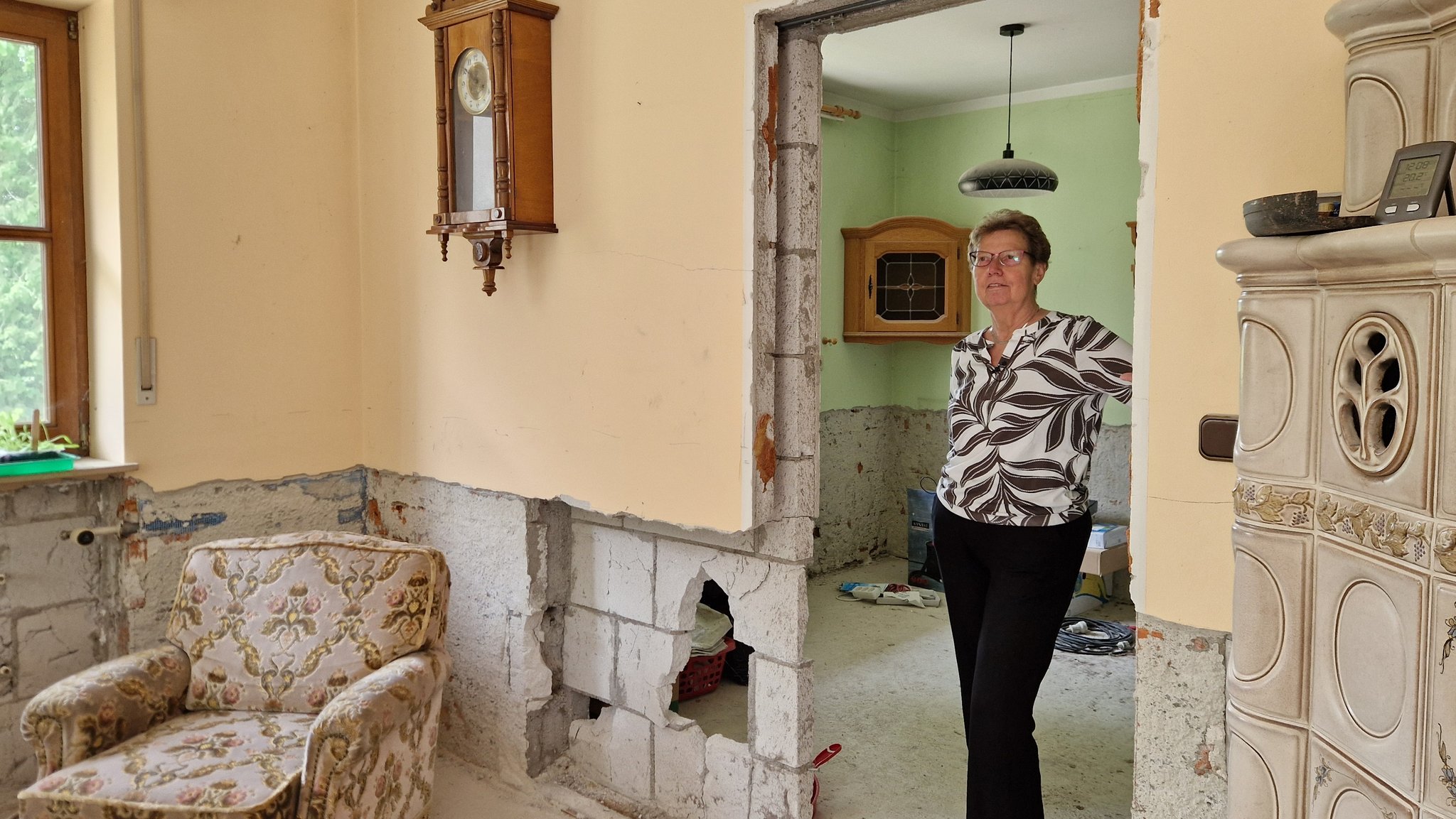 Brigitte Geist steht in ihrem Wohnzimmer: Im unteren Drittel sind die Wände abgetragen bis aufs Mauerwerk, an einer Stelle klafft ein Loch. | Bild: BR/Til Antonie Wiesbeck Brigitte Geist steht in ihrem Wohnzimmer: Im unteren Drittel sind die Wände abgetragen bis aufs Mauerwerk, an einer Stelle klafft ein Loch.