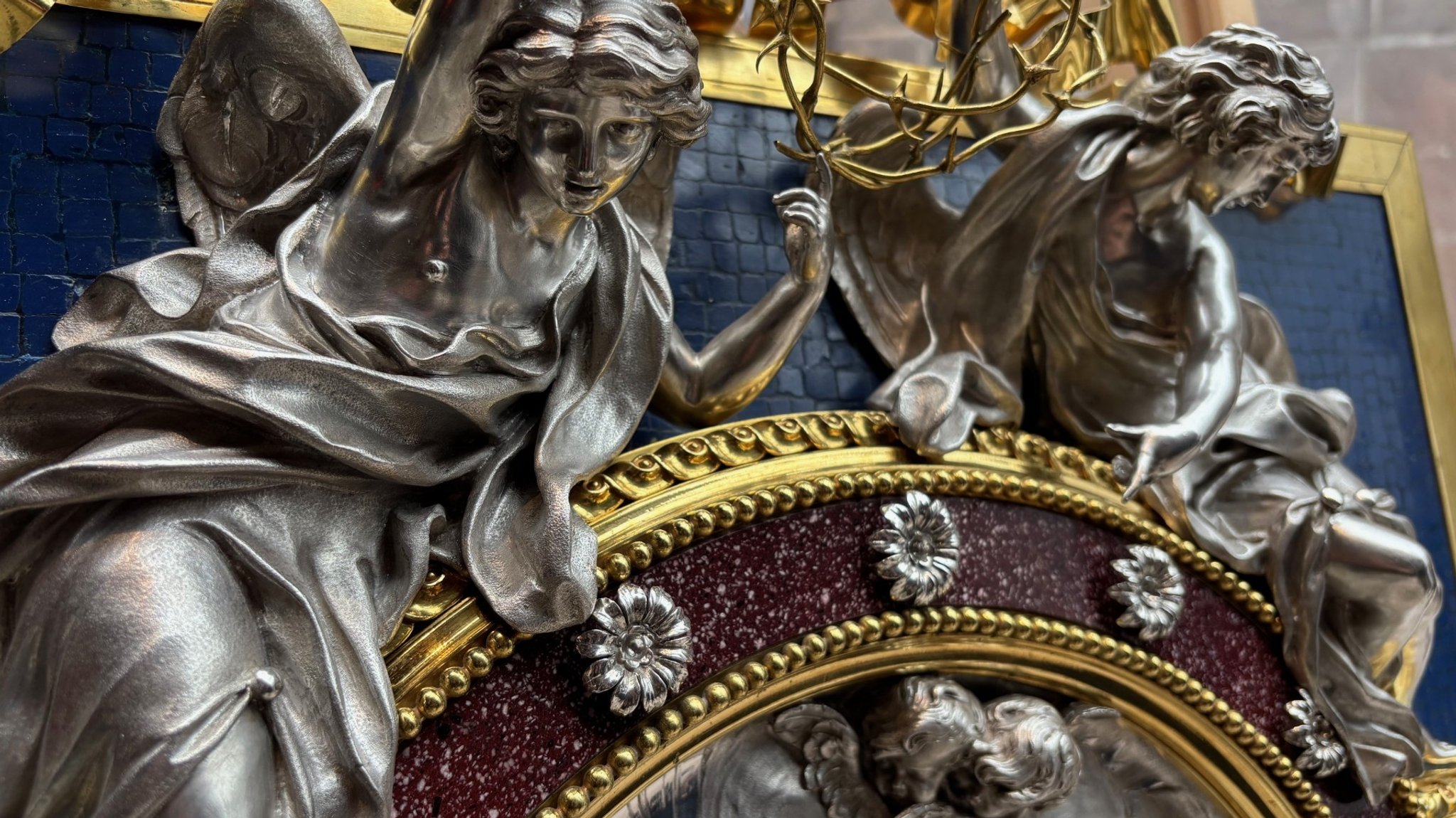 Auf dem Rahmen des Kunstwerks sind zwei silberne Engel zu sehen. 