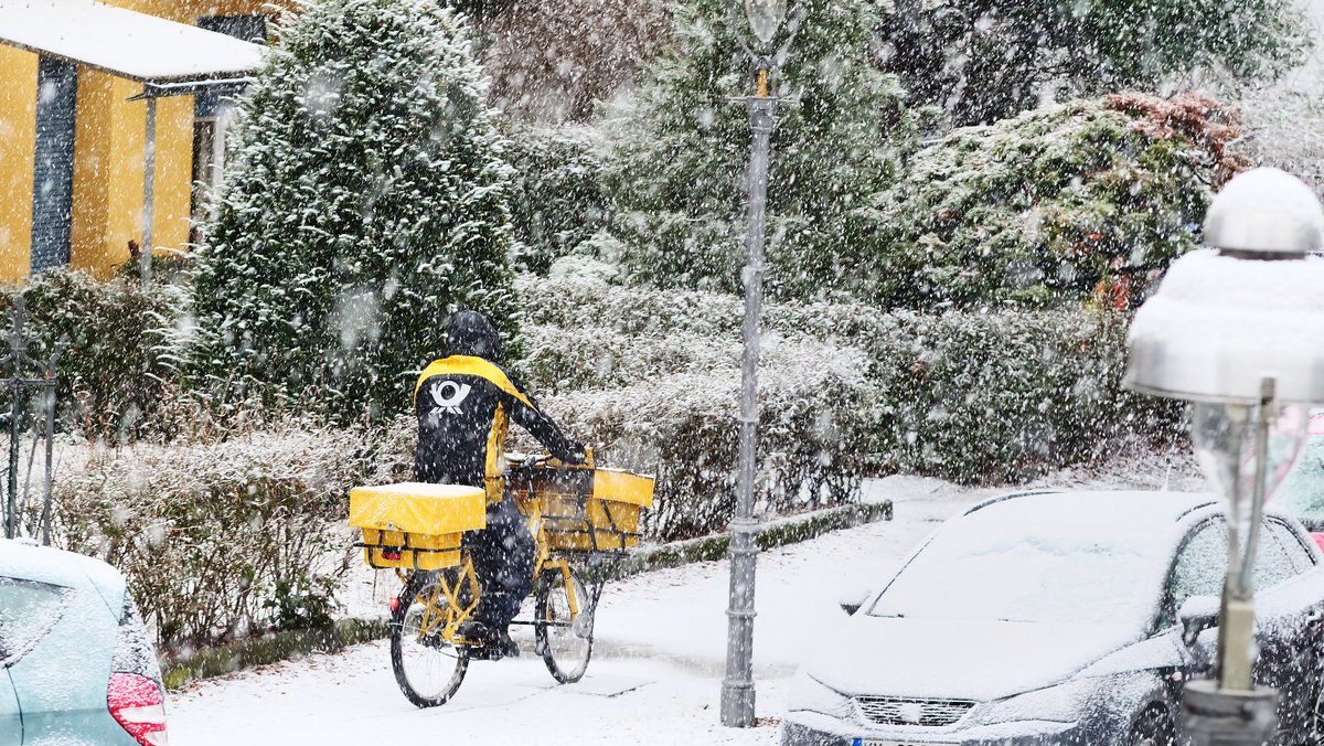 Ein Postbote der Deutschen Post fährt bei Schneefall durch eine Wohnstraße. 