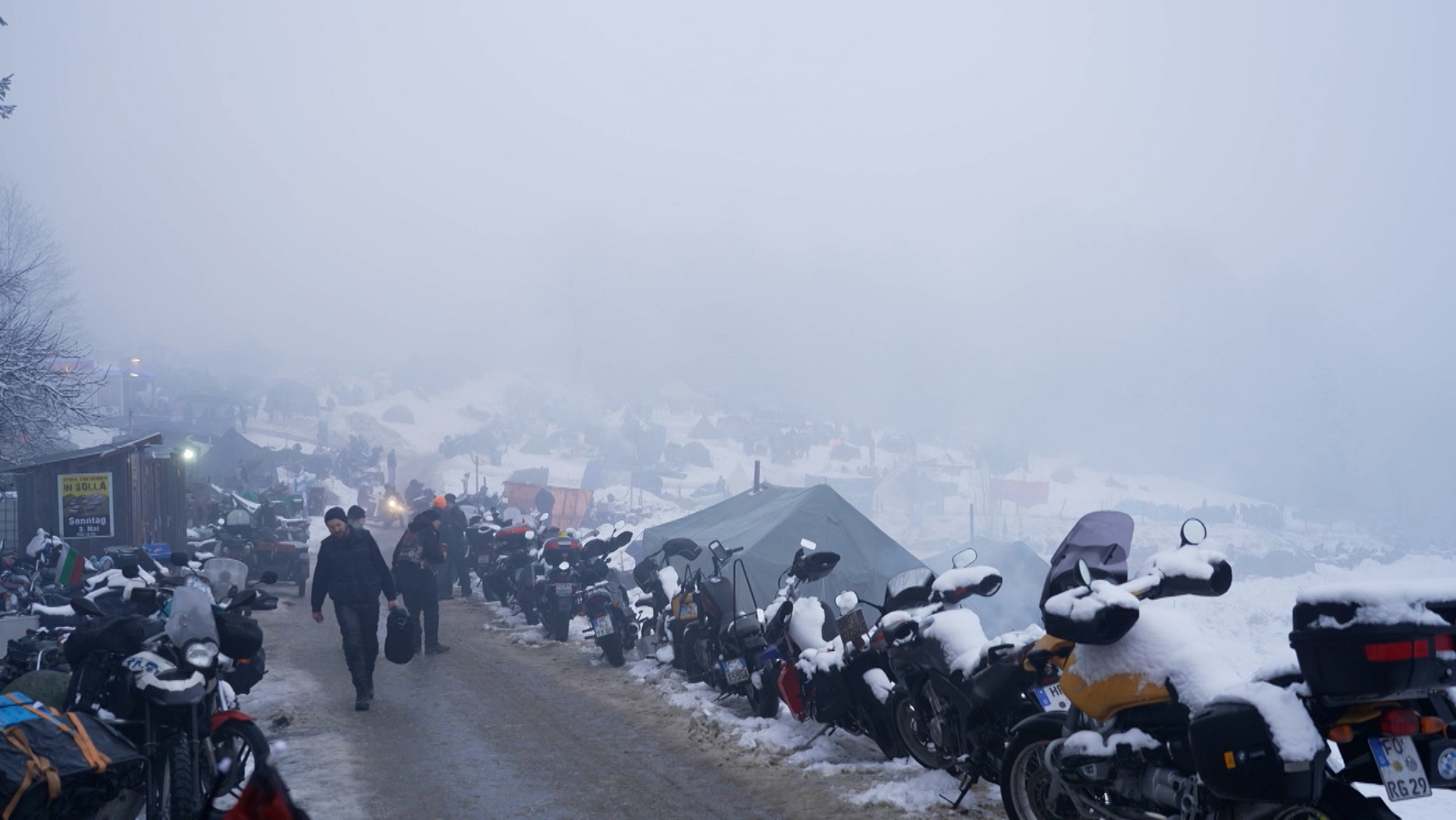 Menschen laufen auf Straße, die seitlich mit verschneiten Motorrädern gesäumt ist | Bild: Bayerischer Rundfunk 2026 Menschen laufen auf Straße, die seitlich mit verschneiten Motorrädern gesäumt ist