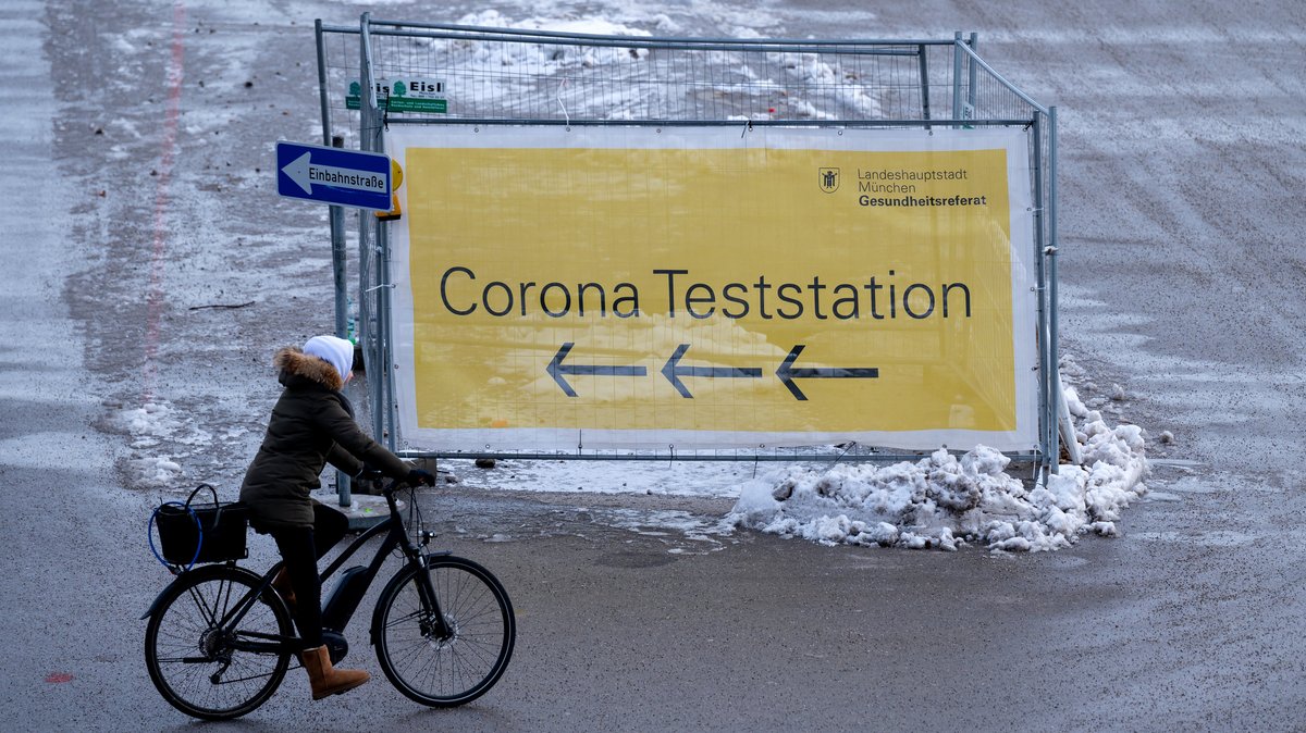 Eine Frau fährt mit ihrem Fahrrad auf der Münchner Theresienwiese an einem Plakat mit der Aufschrift "Corona Teststation" entlang.