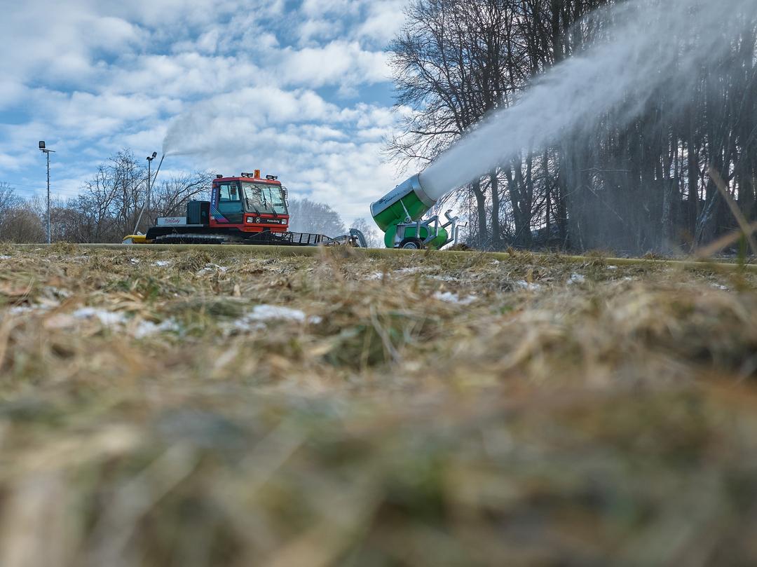 Eine Schneekanone im Einsatz. Außenherum sind kahle Bäume und triste Wiesen zu sehen. Schnee liegt kaum.