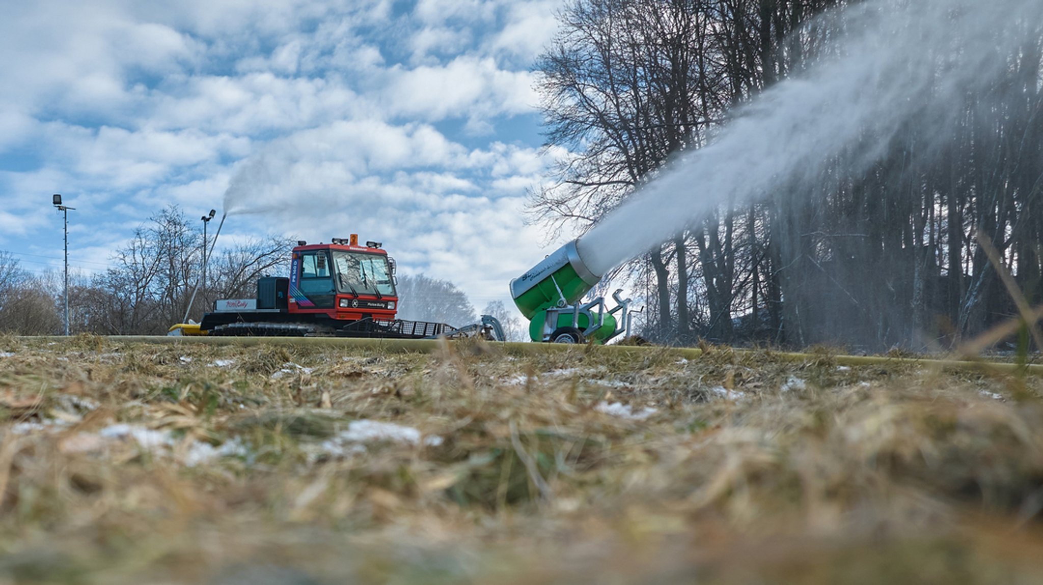 Eine Schneekanone im Einsatz. Außenherum sind kahle Bäume und triste Wiesen zu sehen. Schnee liegt kaum.