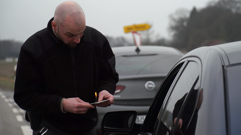 Ein Polizist kontrolliert ein Auto und lässt sich vom Fahrer die Fahrzeugpapiere zeigen. | Bild:BR/Ferdinand Spes