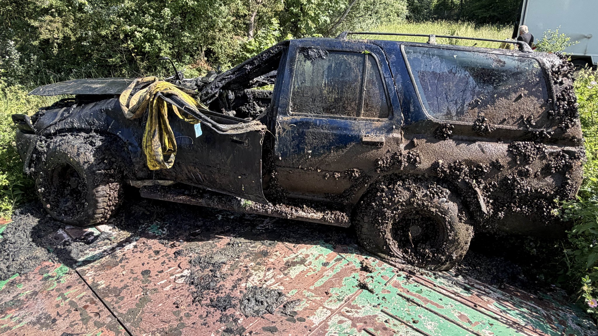 Ein dunkler, demolierter Geländewagen mit platten Reifen ist mit Muscheln übersäht und schlammverschmiert.