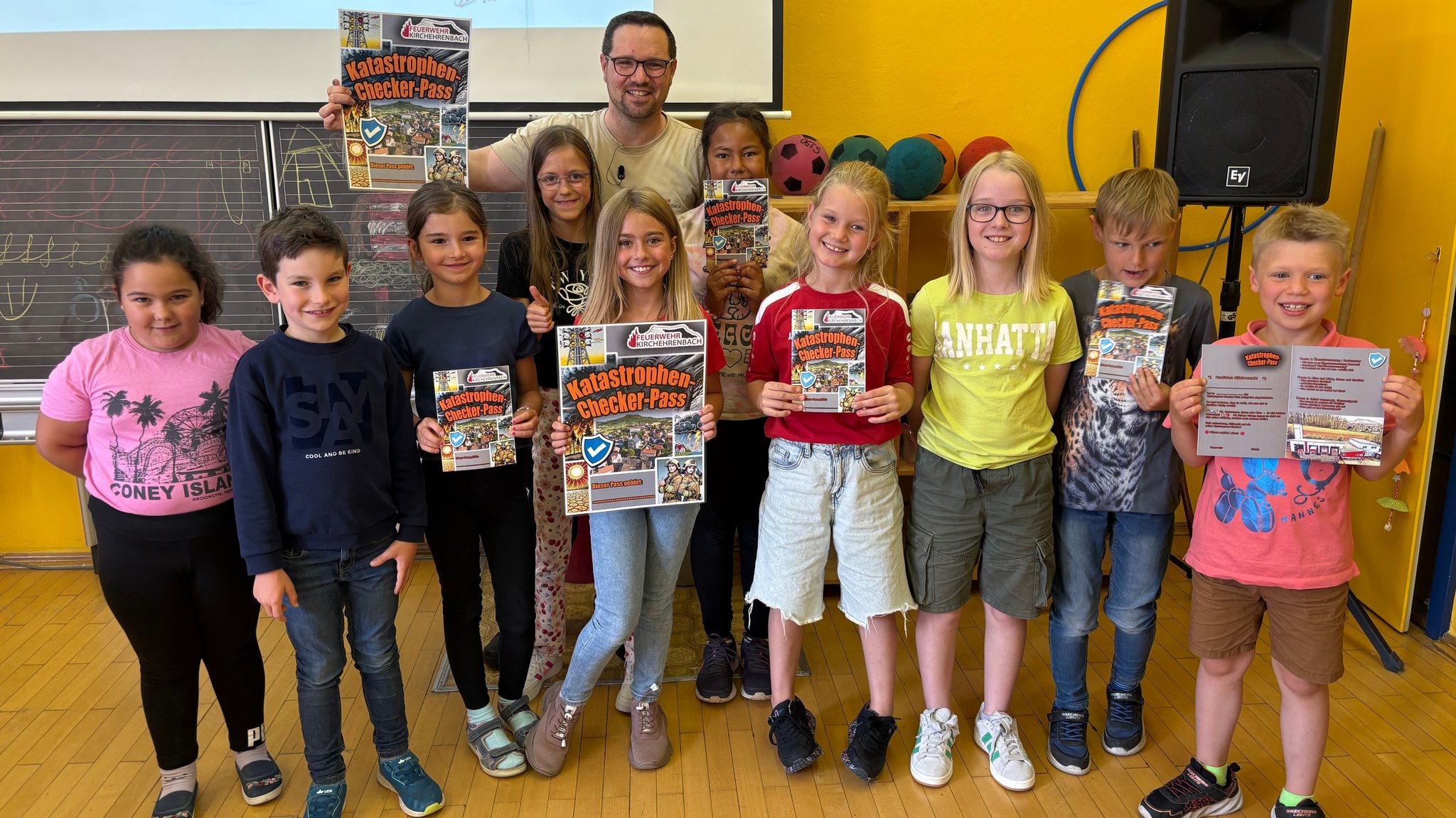Zehn Schulkinder stehen in einem Klassenzimmer und blicken in die Kamera. Einige halten einen Katastrophen-Checker-Pass in den Händen. Hinter ihnen steht ein erwachsener Mann. | Bild: BR/Tobias Burkert Zehn Schulkinder stehen in einem Klassenzimmer und blicken in die Kamera. Einige halten einen Katastrophen-Checker-Pass in den Händen. Hinter ihnen steht ein erwachsener Mann.