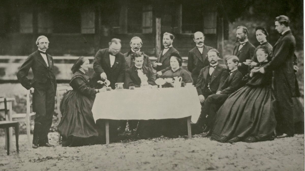 Eine Gruppe von Menschen in Anzug und langen Kleidern beim Picknick vor einer Berghütte mit König Ludwig II. 