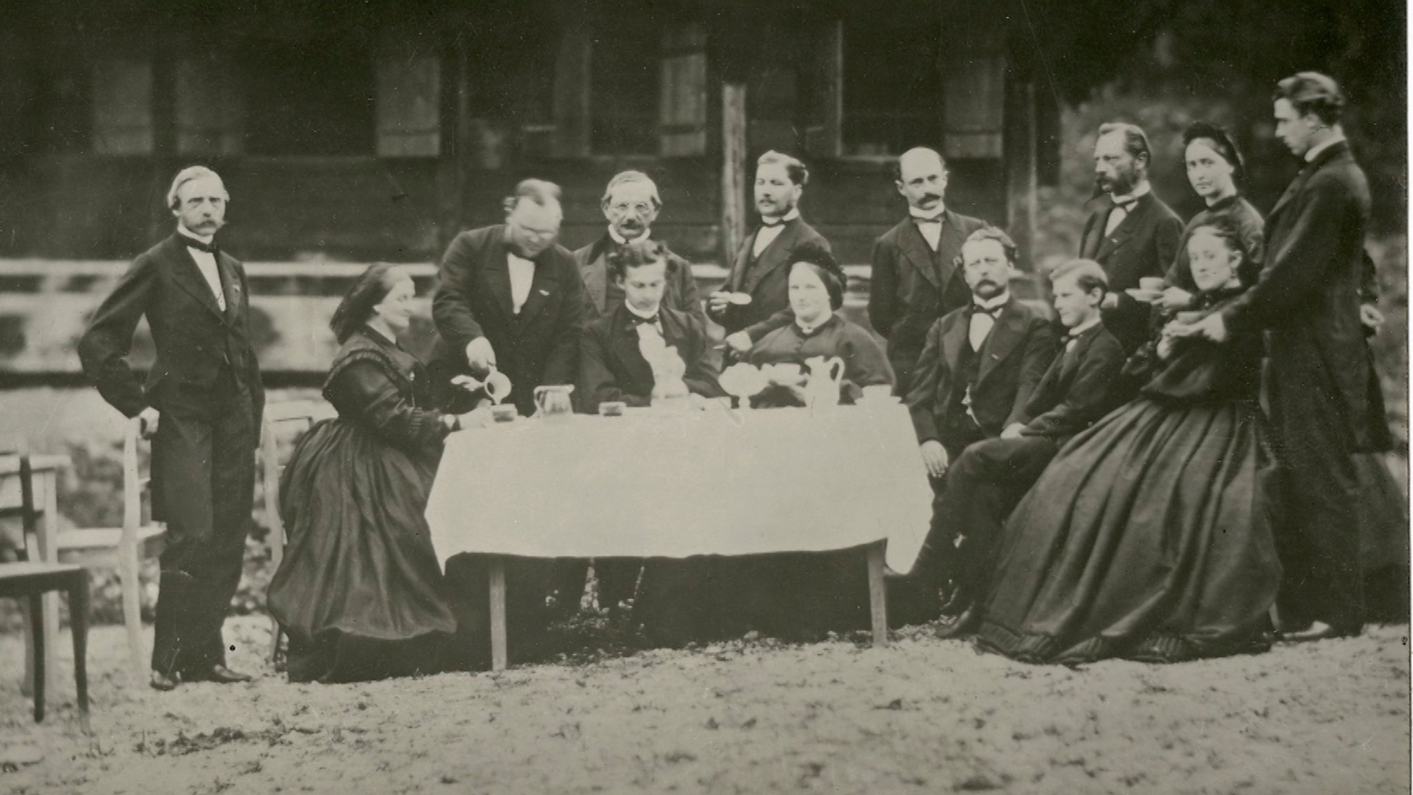 Eine Gruppe von Menschen in Anzug und langen Kleidern beim Picknick vor einer Berghütte mit König Ludwig II. 