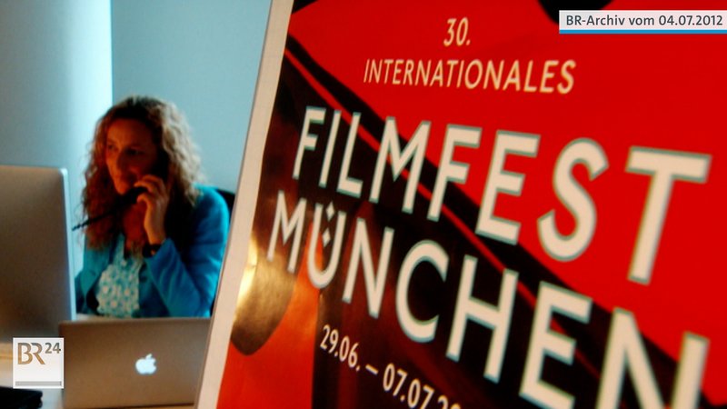Links  Diana Iljine vor einem Computer sitzend, telefonierend, rechts Plakat zum 30. Filmfest München