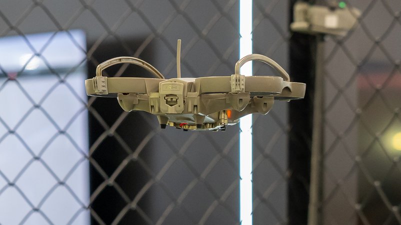 Eine taktische Überwachungsdrohne schwebt in einem abgesicherten Testbereich mit Sicherheitsnetz. Die kompakte Drohne ist speziell für militärische und polizeiliche Anwendungen konzipiert und verfügt über Schutzbügel um die Rotoren. Im Hintergrund sind Bildschirme und Beleuchtungselemente zu erkennen | Bild: picture alliance / Eibner-Pressefoto | Eibner-Pressefoto/Ardan Fuessman Eine taktische Überwachungsdrohne schwebt in einem abgesicherten Testbereich mit Sicherheitsnetz. Die kompakte Drohne ist speziell für militärische und polizeiliche Anwendungen konzipiert und verfügt über Schutzbügel um die Rotoren. Im Hintergrund sind Bildschirme und Beleuchtungselemente zu erkennen