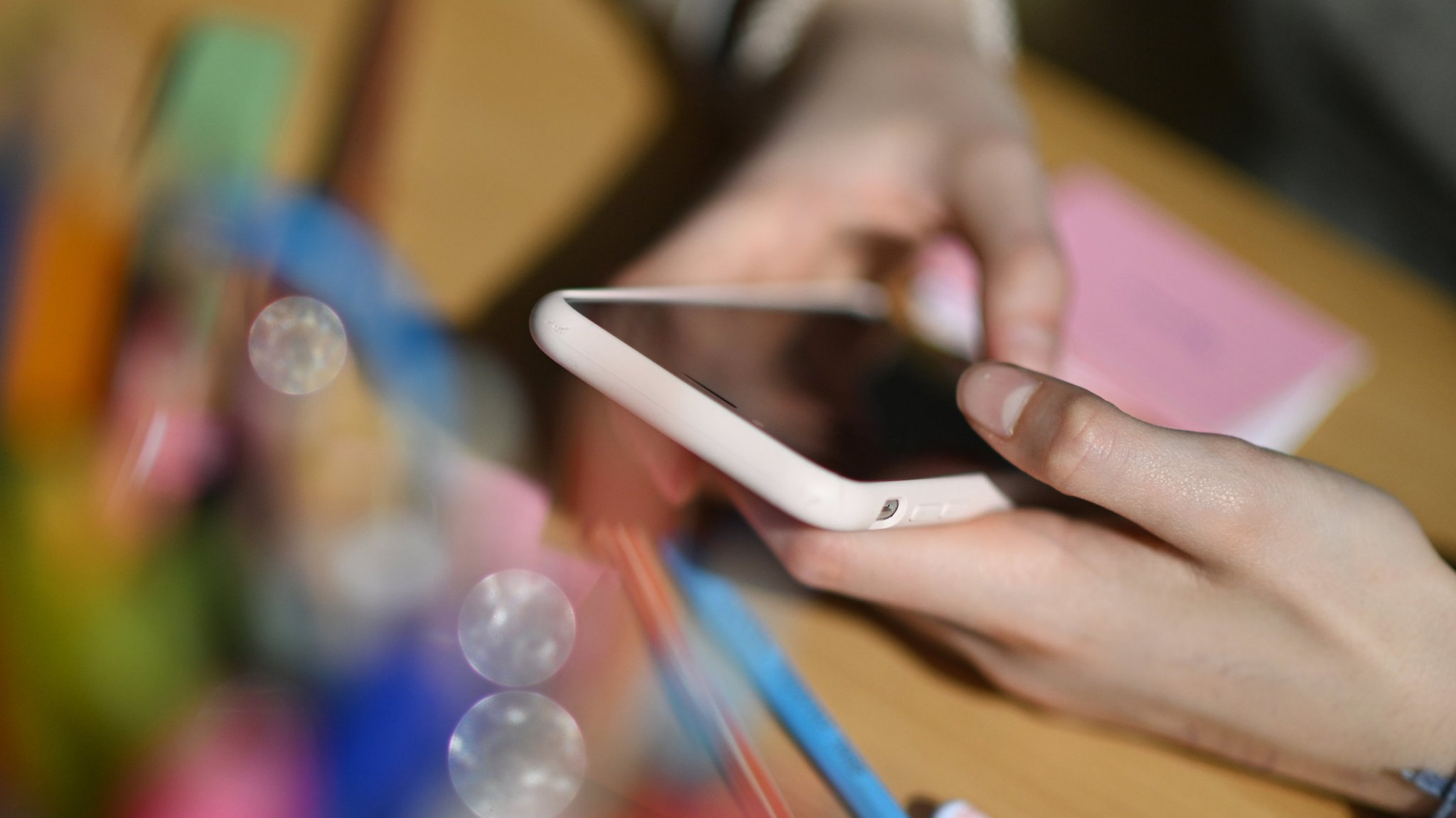 Ein Schulkind sitzt an einem Schreibtisch mit Schulmaterialien und hält ein Smartphone in den Händen.