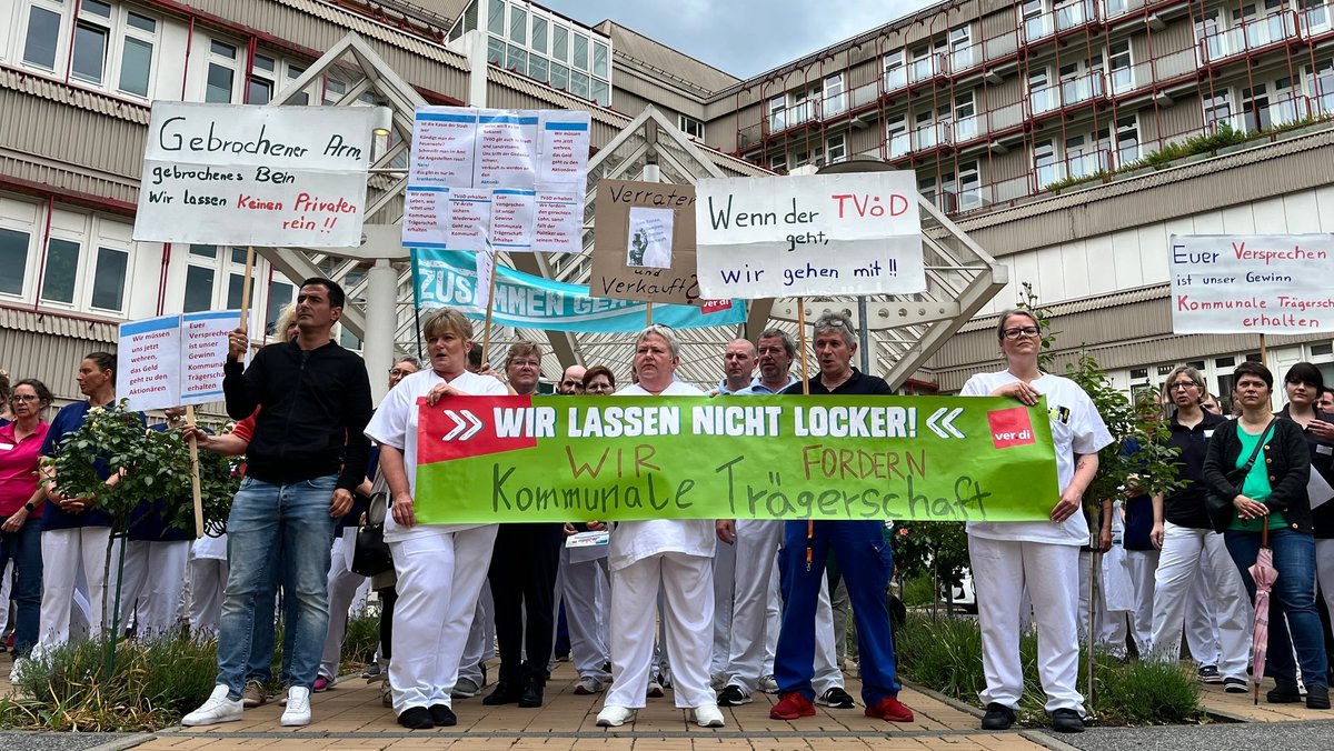 Krankenhaus-Mitarbeiter halten Transparente in die Höhe, auf denen steht unter anderem: "Wir lassen nicht locker, wir fordern kommunale Trägerschaft". 