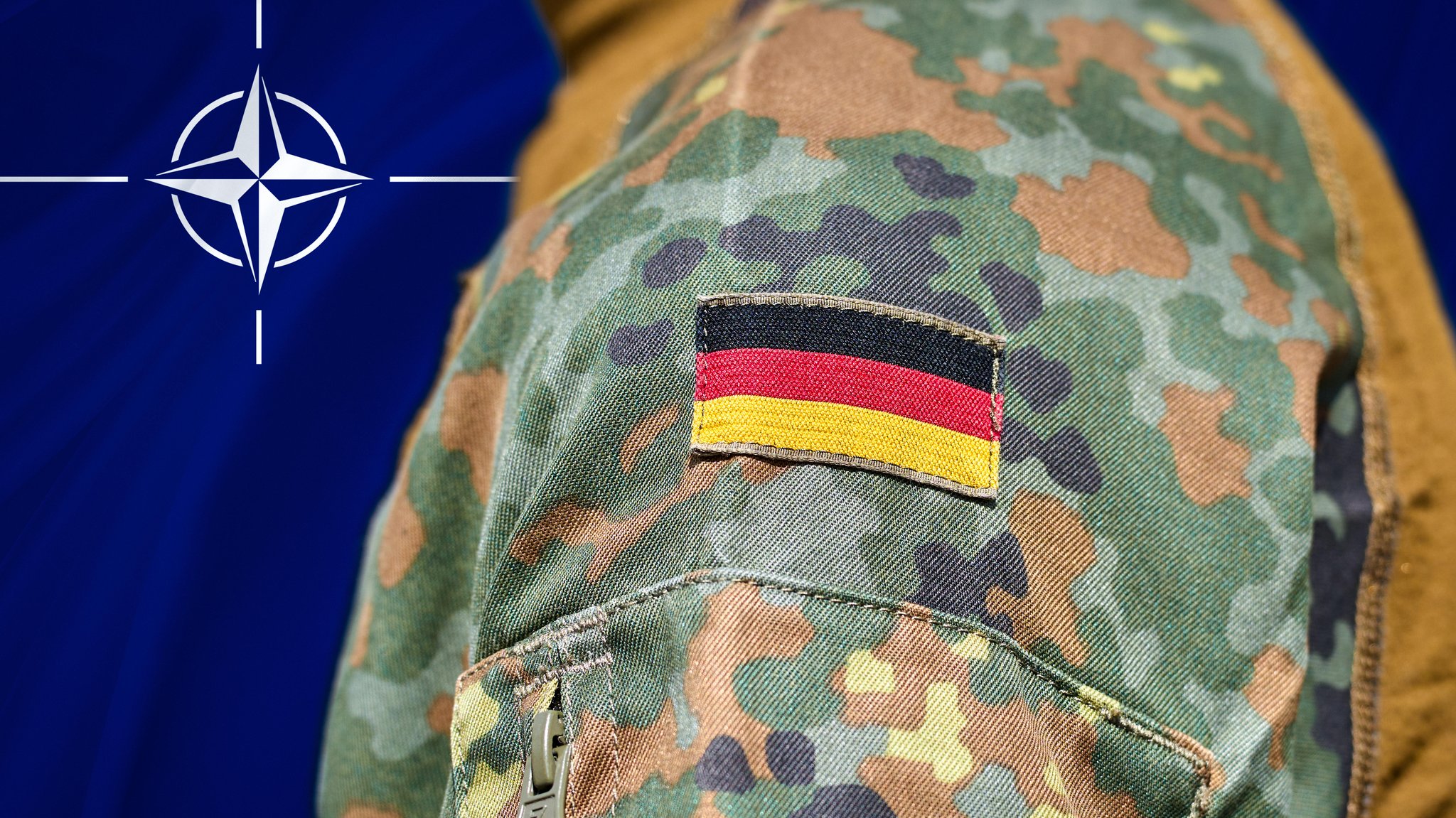 Ein Soldat in deutscher Tarnuniform mit aufgenähter Deutschlandflagge steht vor einer Nato-Flagge.