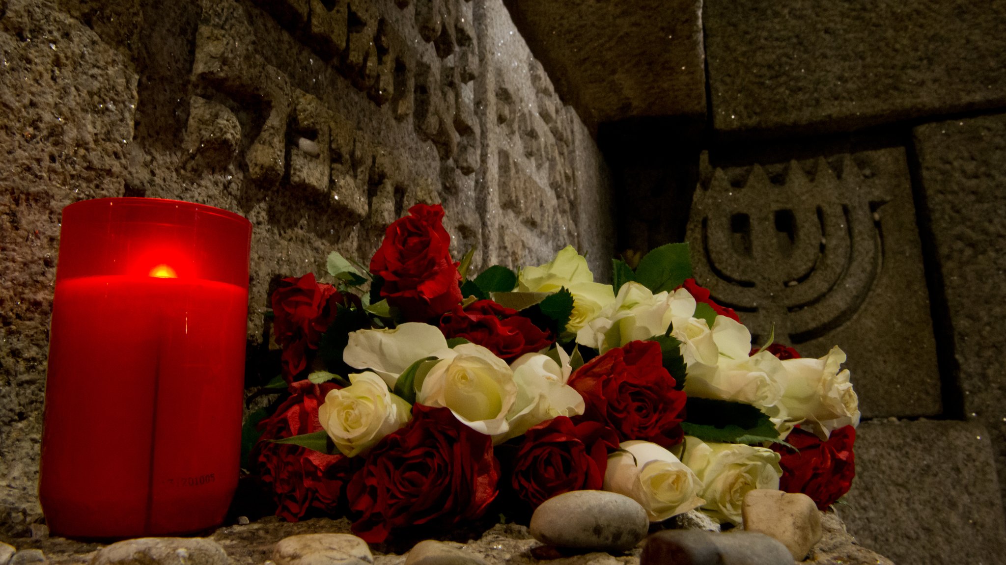 Blumen und eine Friedhofskerze auf dem Gedenkstein, der an der Stelle der ehemaligen Hauptsynagoge in München steht. Im Hintergrund der siebenarmige Leuchter in Stein gehauen.    