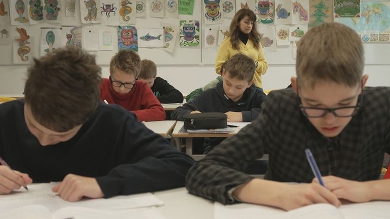 In einer Schulklasse sitzen Jugendliche beim Schreiben eines Texts am Schreibtisch. Im Hintergrund steht die Lehrerin der Brückenklasse an der Realschule Freiham. 