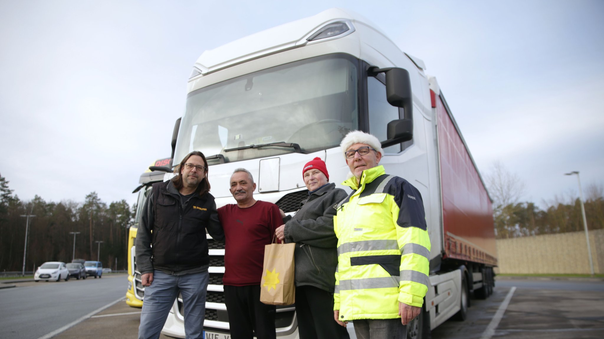 Jens Harzbecker, Anita Schubert und Klaus Elmer stehen mit einem Trucker auf einem Rastplatz und übergeben eine Tüte.