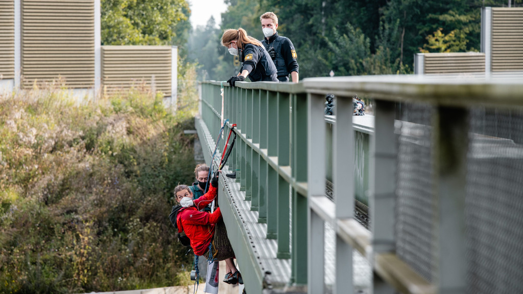 Klimaaktivisten seilen sich an einer Brücke ab. Oben an der Brücke stehen eine Polizistin, die sich zu den Aktivisten herunterbeugt, und ein Polizist.
