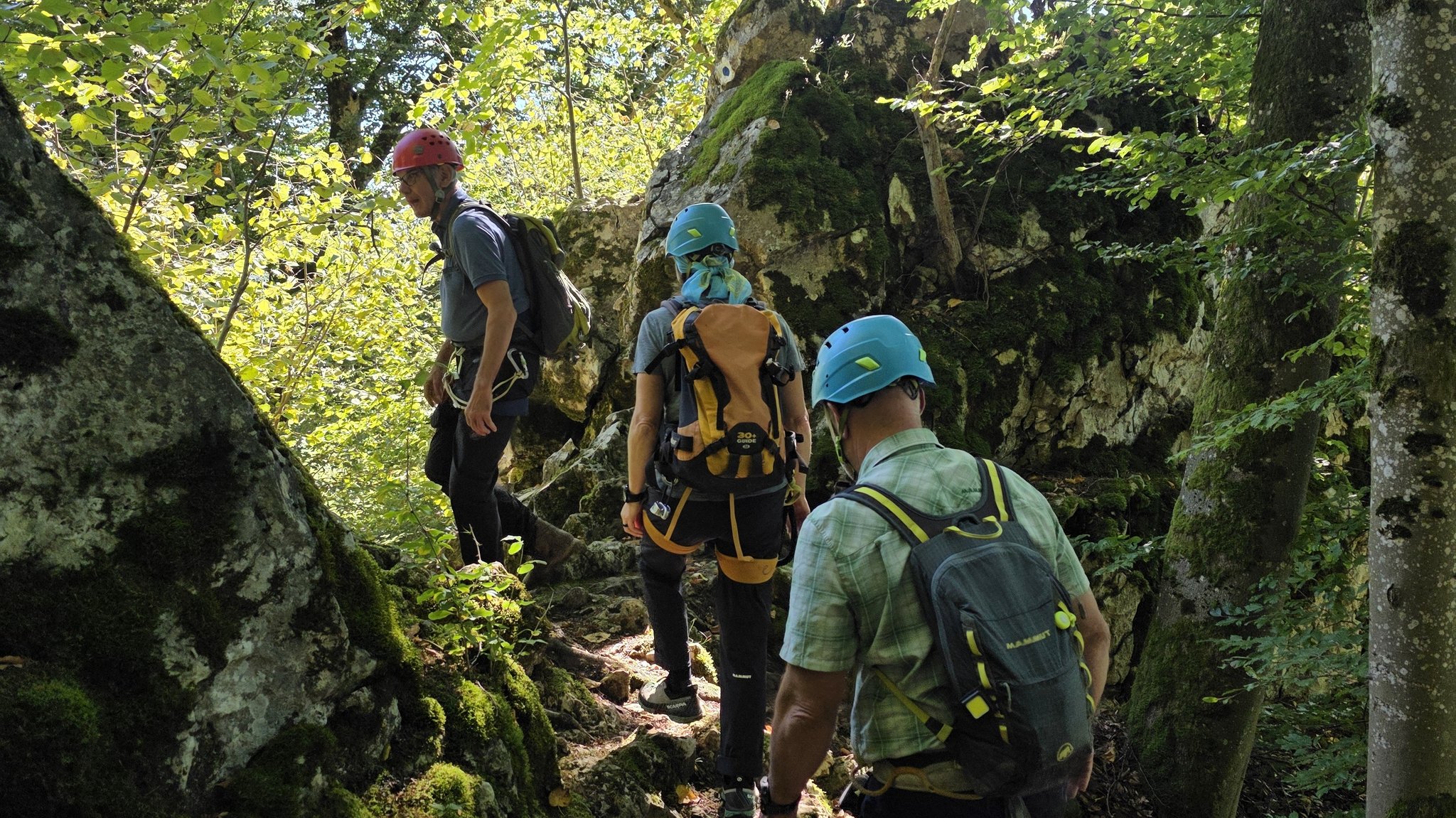 Drei Wandernde schreiten mit Helmen und Rucksack über einen felsigen Steig.