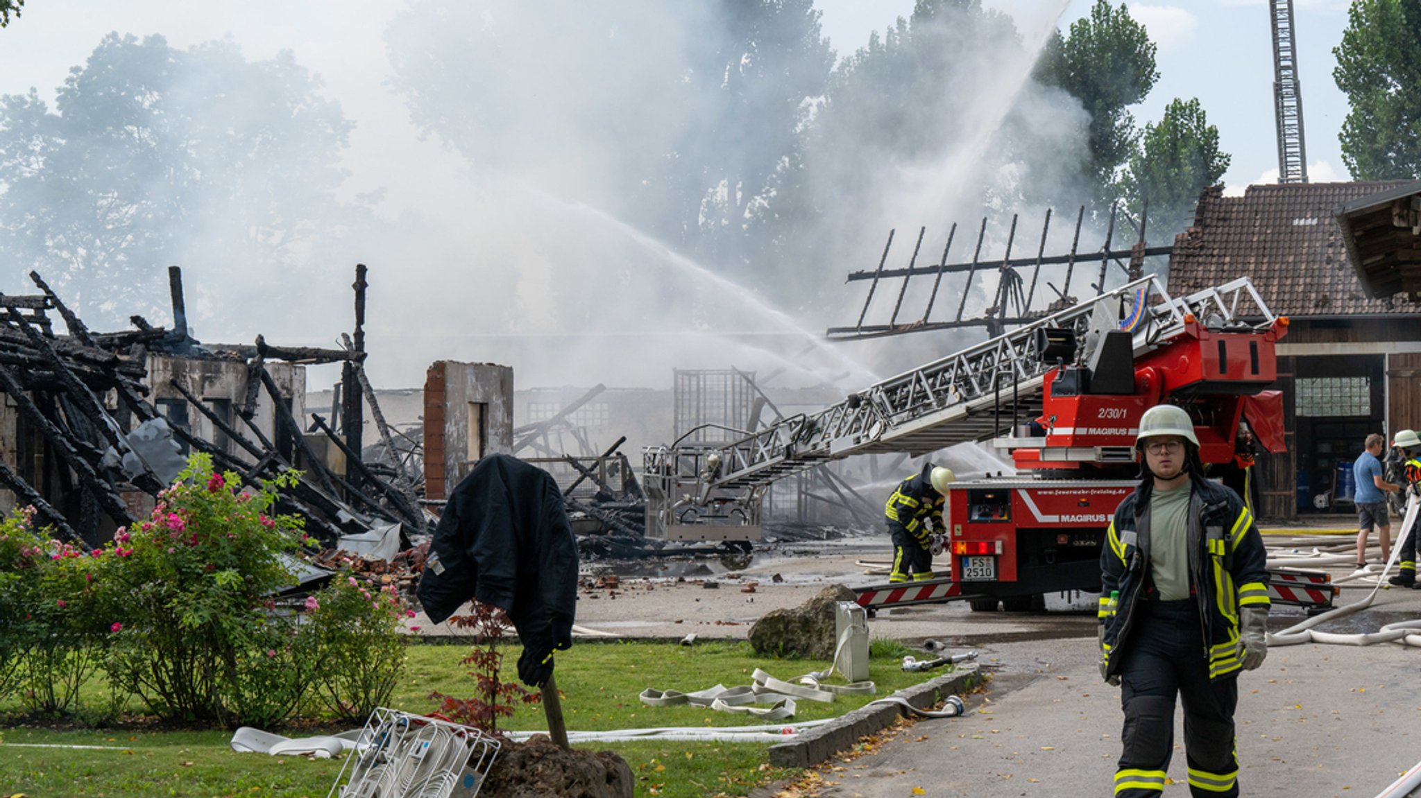 Feuerwehrleute löschen in einem landwirtschaftlichen Betrieb. Im Hintergrund ein völlig abgebrannter Stall.