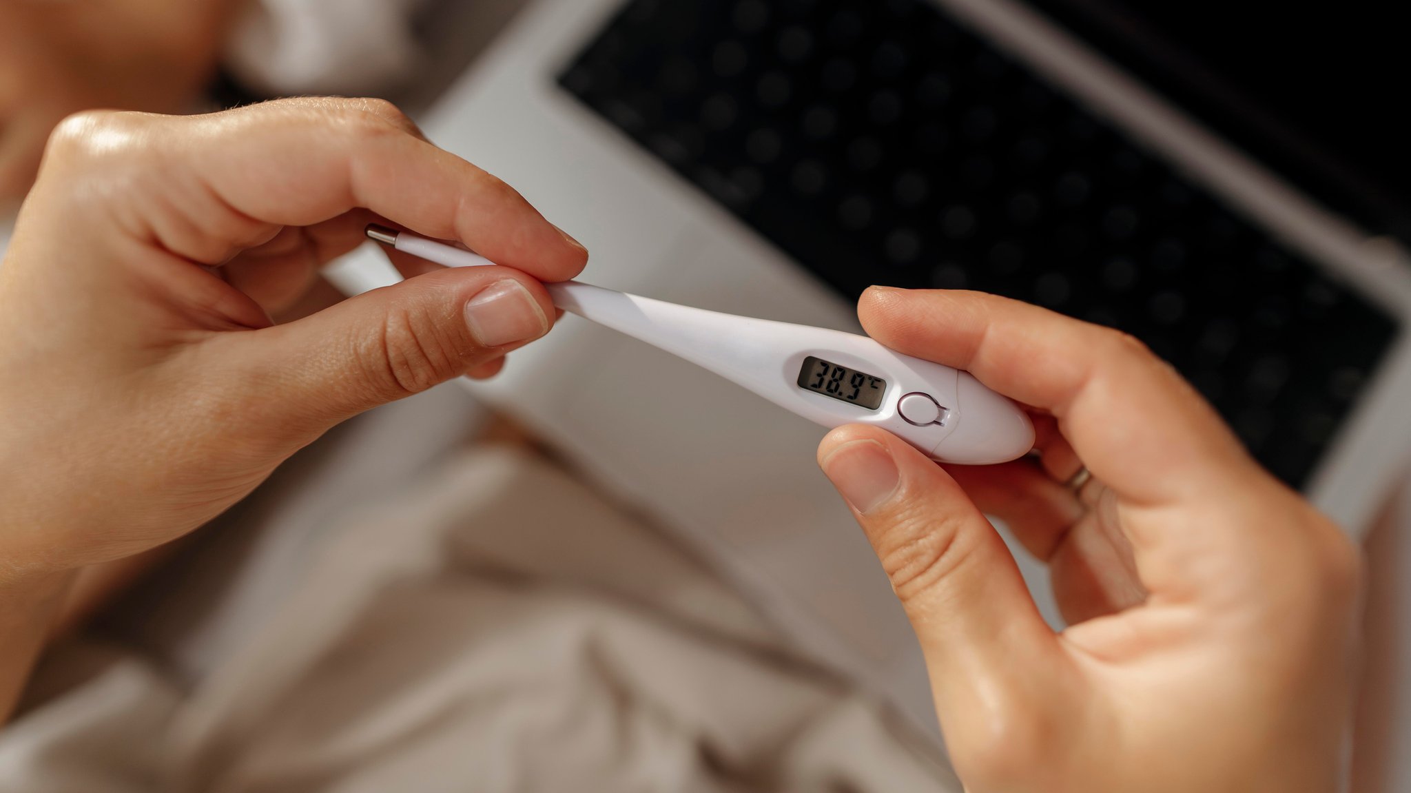 Hände, die ein Fieberthermometer halten, es zeigt eine Temperatur von 38,9 an.  