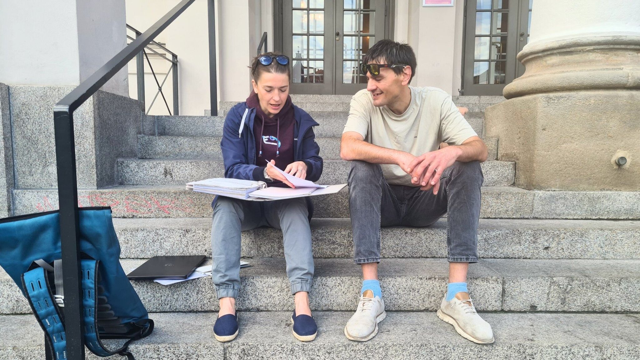 Martina und Danijel sitzen auf den Stufen und blättern durch einen Aktenordner | Bild: BR/Birgit Rätsch Martina und Danijel sitzen auf den Stufen und blättern durch einen Aktenordner